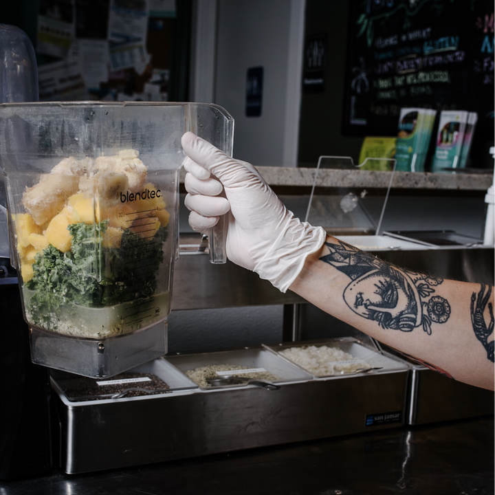 A tattooed arm operates a blender full of fruits and vegetables
